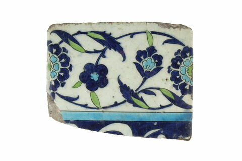 Carreau de bordure fragmentaire à inscription (?) sur fond bleu sombre. Bordure à rinceaux fleuris de rosettes et fleurettes